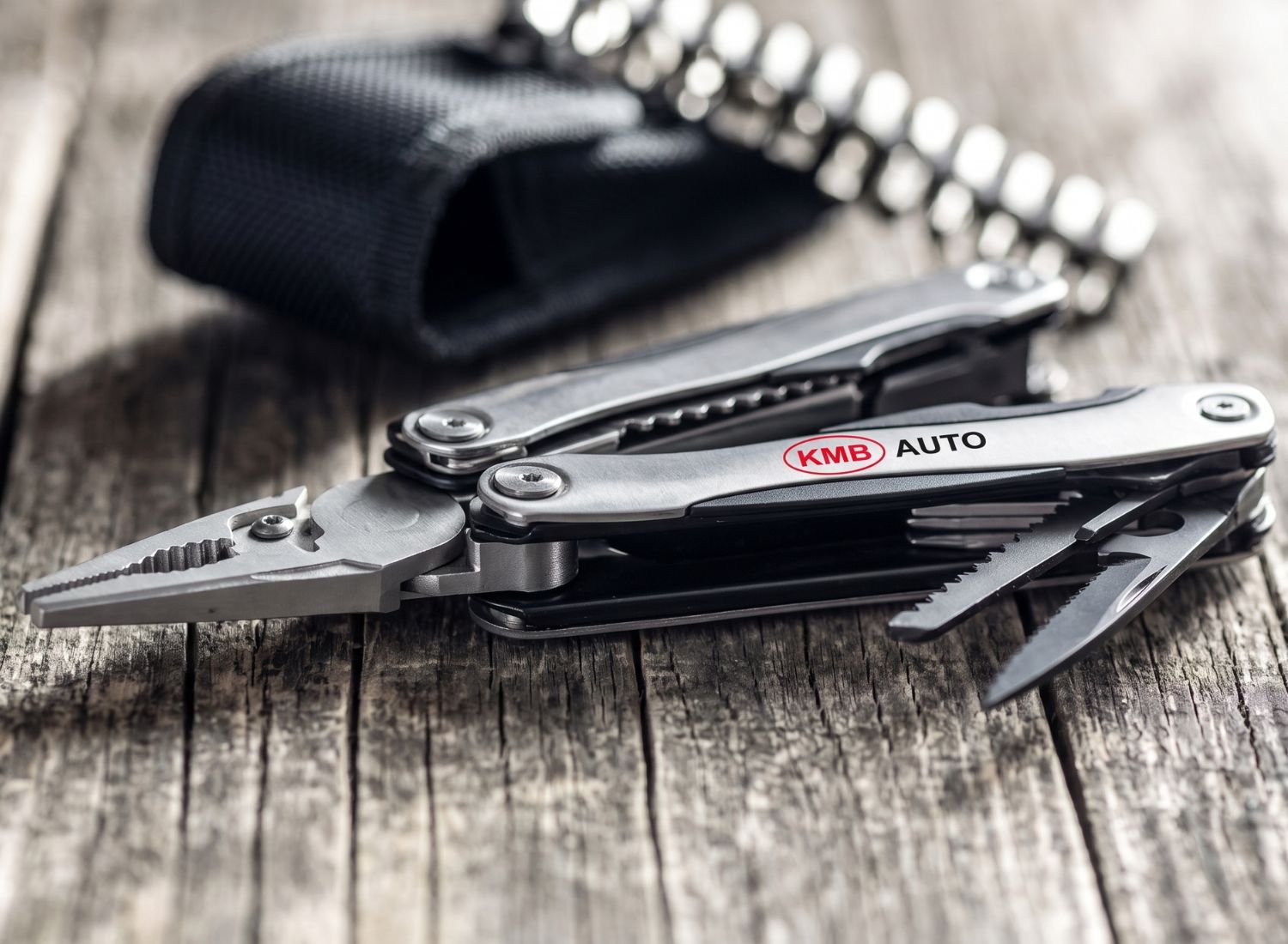 Stalowy multitool reklamowy ze szczypcami i grawerowanym logo KMB AUTO obok czarnego etui. Praktyczne gadżety dla inżynierów.