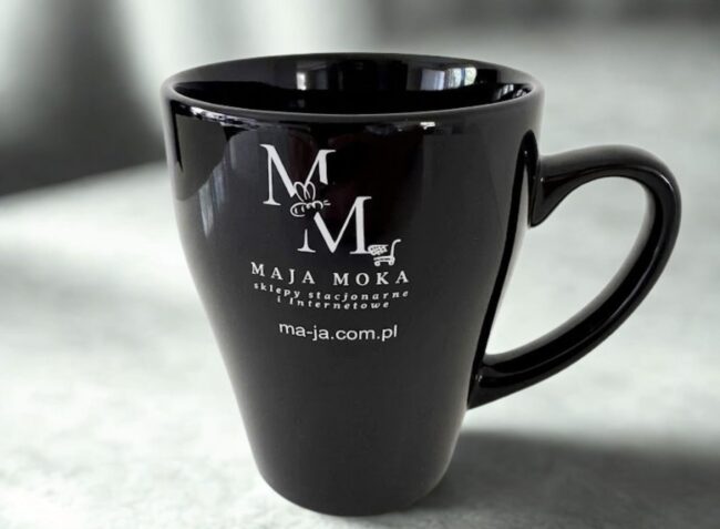 Czarny, lśniący kubek ceramiczny z białym, kontrastowym nadrukiem logo MAJA MOKA. Eleganckie upominki dla klientów.