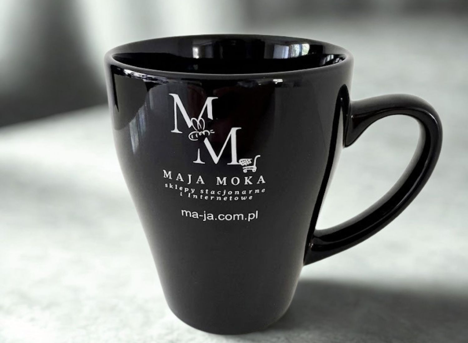 Czarny, lśniący kubek ceramiczny z białym, kontrastowym nadrukiem logo MAJA MOKA. Eleganckie upominki dla klientów.