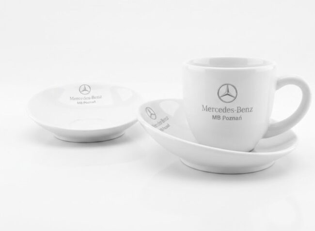 Elegancka biała filiżanka ze spodkiem i szarym logo Mercedes-Benz. Prestiżowa ceramika reklamowa dla dealerów samochodowych.