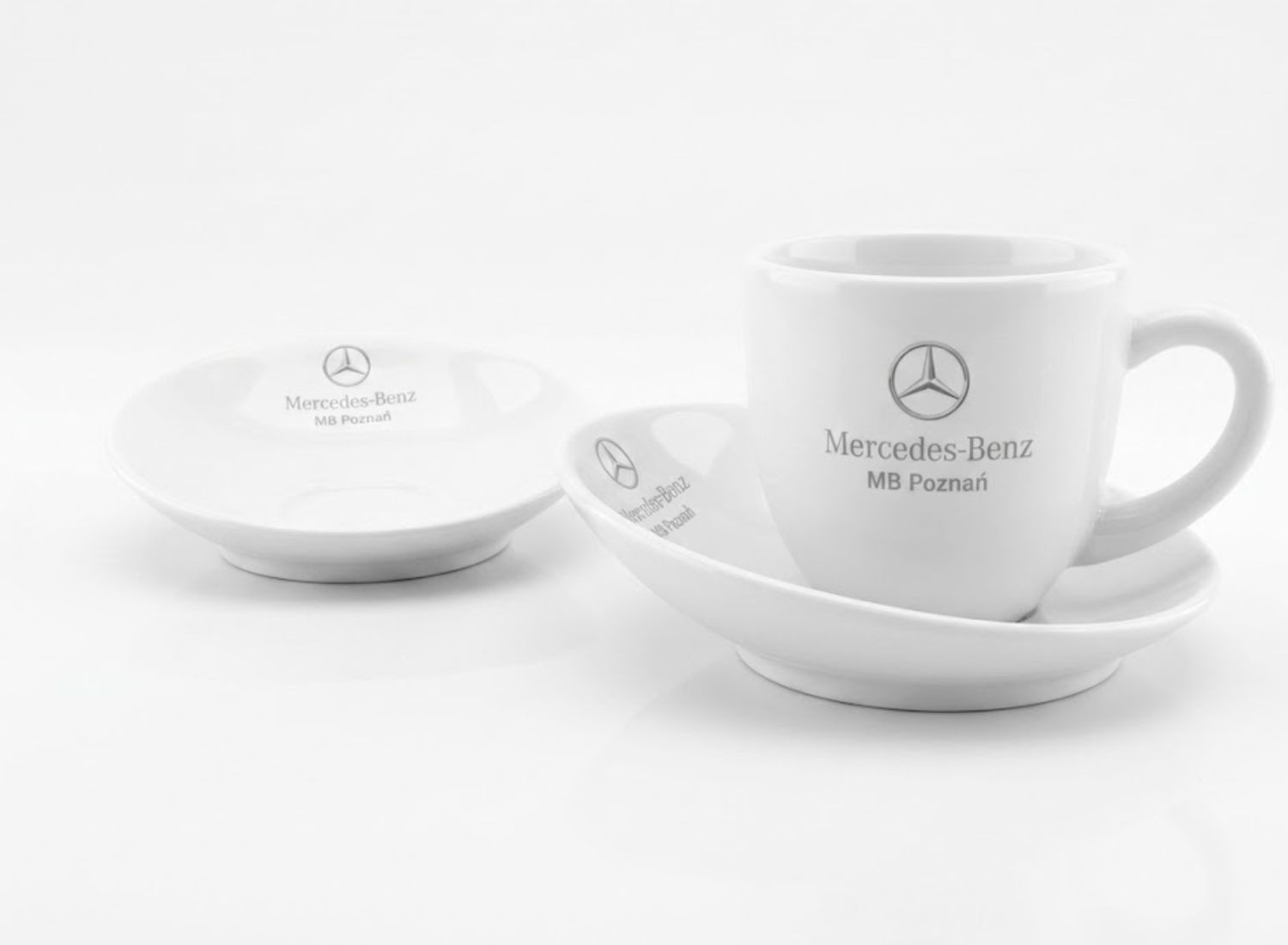 Elegancka biała filiżanka ze spodkiem i szarym logo Mercedes-Benz. Prestiżowa ceramika reklamowa dla dealerów samochodowych.