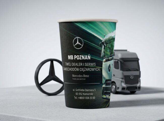 Papierowy kubek ekologiczny z logotypem i grafiką pojazdu ciężarowego Mercedes-Benz obok emblematu na biurku.