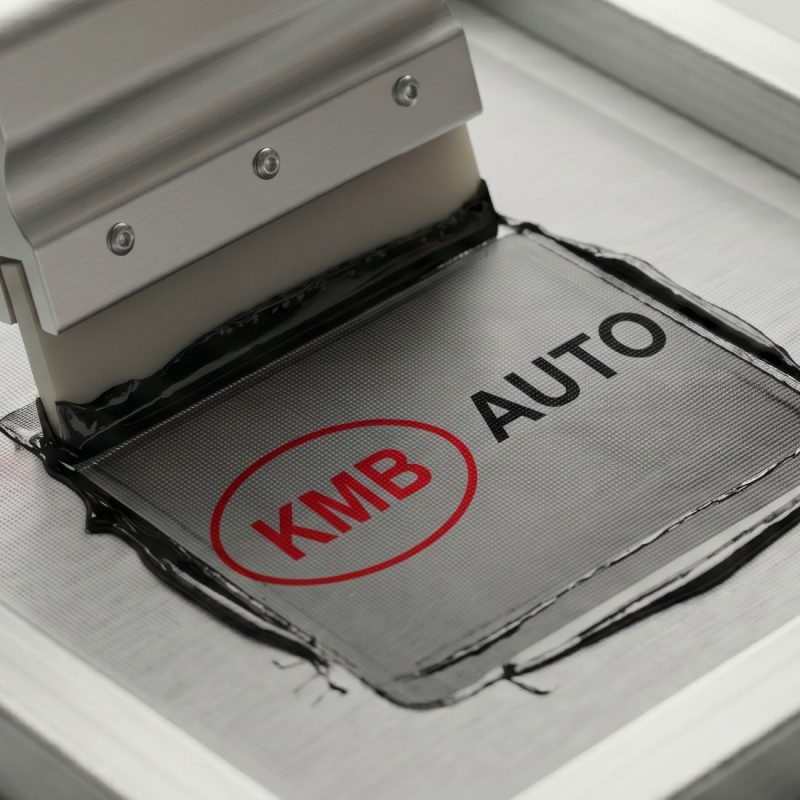 Maszyna do sitodruku przeciskająca czarną i czerwoną farbę przez matrycę z logo KMB AUTO. Trwałe znakowanie odzieży roboczej i toreb.