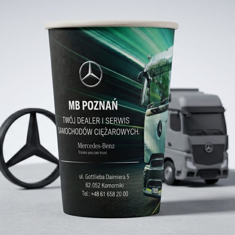 Papierowy kubek ekologiczny z logotypem i grafiką pojazdu ciężarowego Mercedes-Benz obok emblematu na biurku.